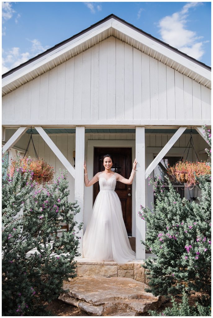 Bride in long sleeve flowy white wedding gown poses in front of white cottage bridal suite at Stonebridge wedding and event venue photographed by Dallas wedding photographer Picture Bouquet Studio. 