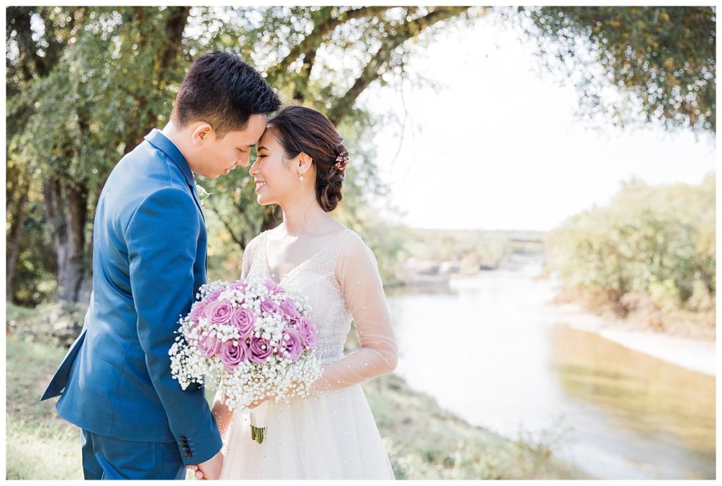 Bride in long sleeve white wedding dress and groom in navy blue suit poses in front of water for portrait session for outdoor wedding at Stonebridge Wedding Venue photographed by Dallas wedding photographer Picture Bouquet Studio. 