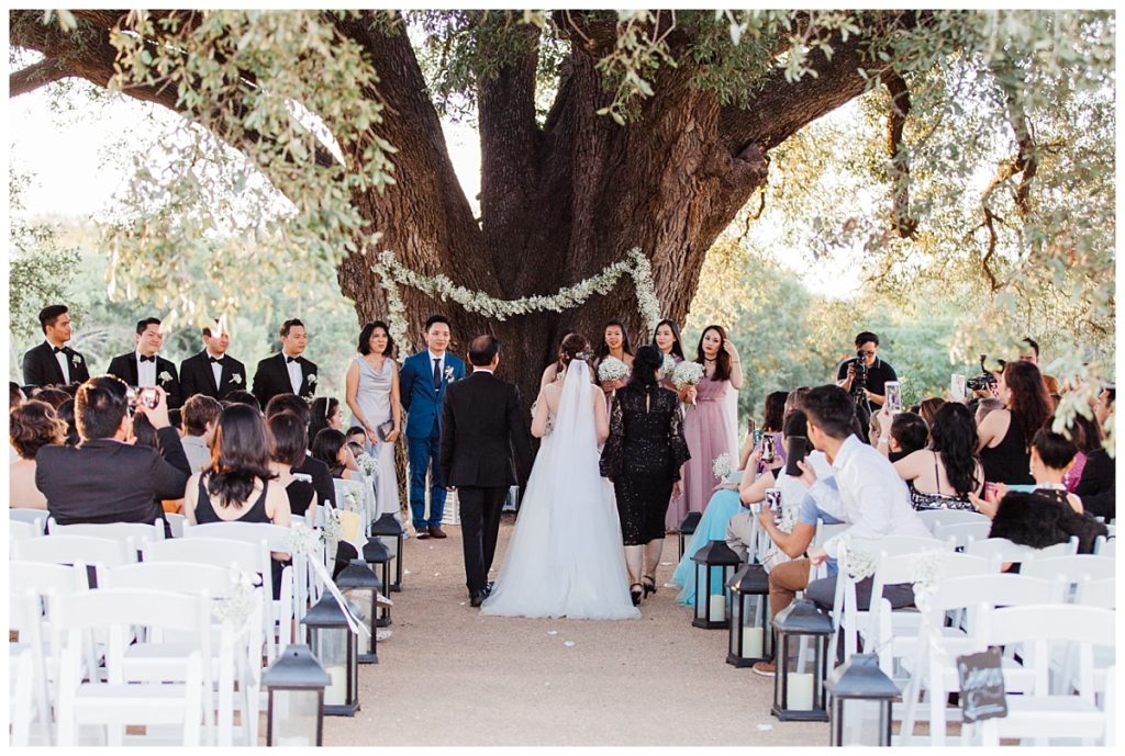 Shot of bride and parents walking down the aisle towards groom in front of oak tree for outdoor wedding at Stonebridge Wedding Venue photographed by Dallas wedding photographer Picture Bouquet Studio. 