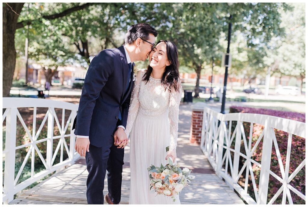 Bride in elegant, lace, minimal white dress shares laugh with groom in navy suit on bridge at Haggard Park in Plano, TX for bridal party portraits by wedding photographer Jenny Bui of Picture Bouquet Studio, a Dallas based wedding photography studio. 