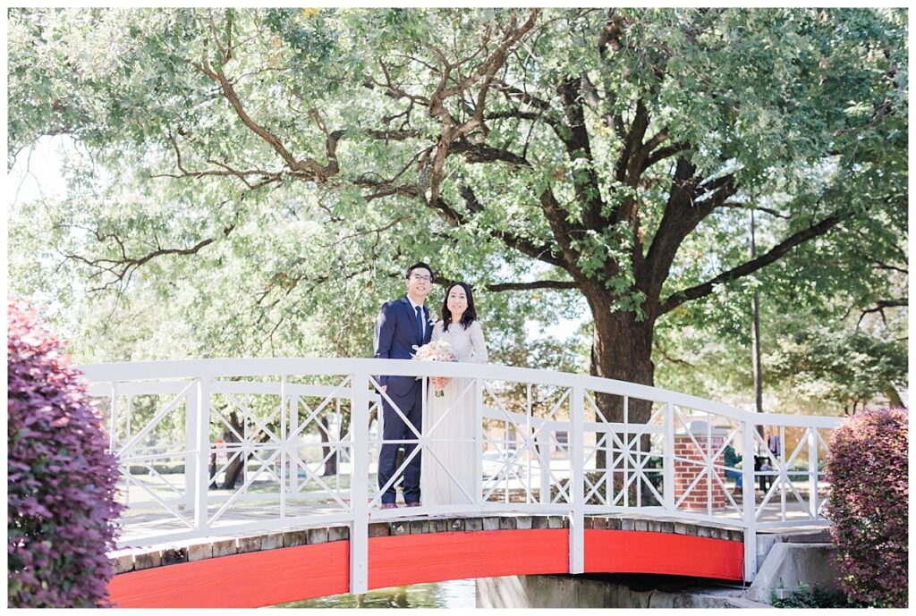 Bride in elegant, lace, minimal white dress poses with groom in navy suit on bridge at Haggard Park in Plano, TX for bridal party portraits by wedding photographer Jenny Bui of Picture Bouquet Studio, a Dallas based wedding photography studio. 