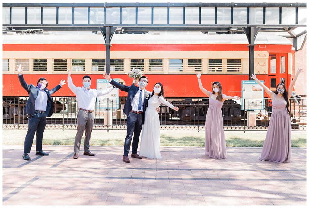 Bride in elegant, lace, white minimal dress holding soft orange bouquet and groom in navy suit poses with bridal party in front of red train at Haggard Park in Plano, TX for bridal party portraits by wedding photographer Jenny Bui of Picture Bouquet Studio, a Dallas based wedding photography studio. 
