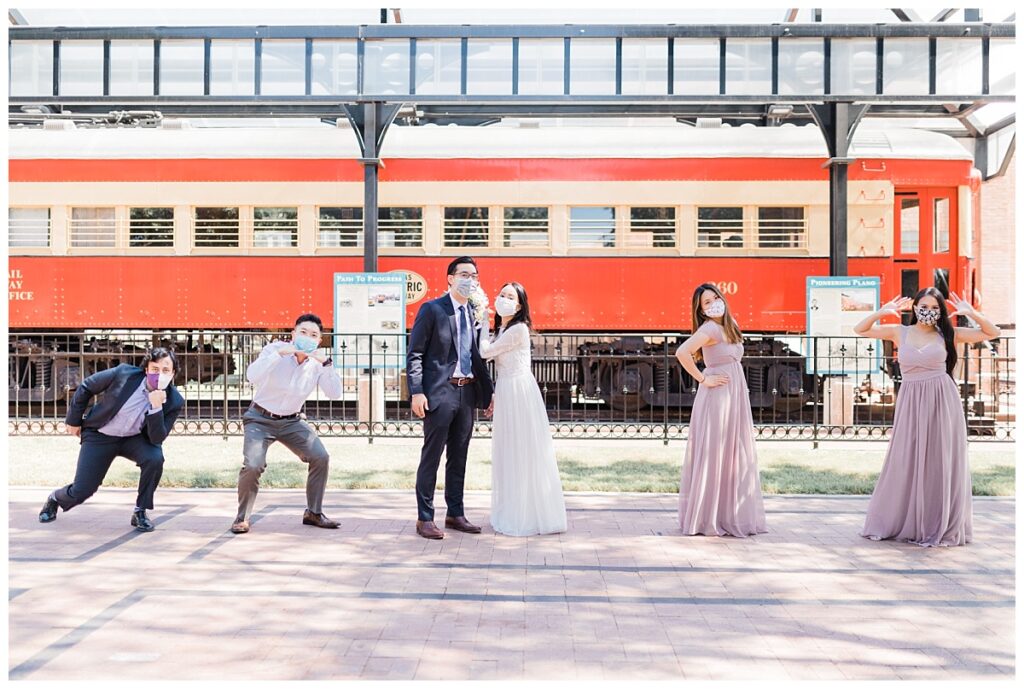 Bride in elegant, lace, white minimal dress holding soft orange bouquet and groom in navy suit poses with bridal party in front of red train at Haggard Park in Plano, TX for bridal party portraits by wedding photographer Jenny Bui of Picture Bouquet Studio, a Dallas based wedding photography studio. 