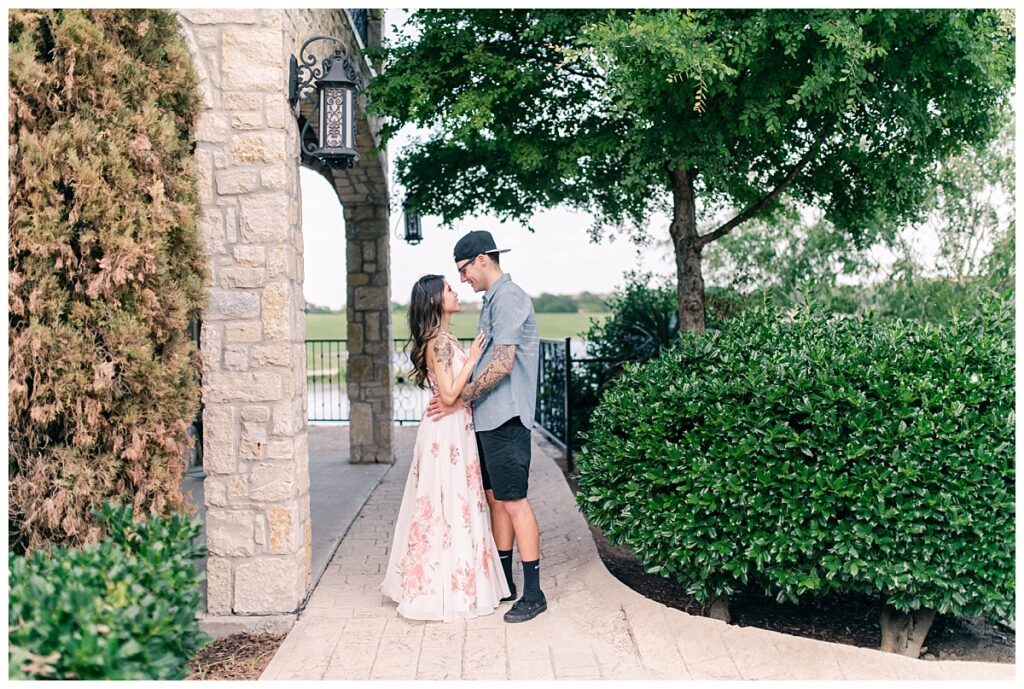 Women in long flowy white dress holds fiance along pathway for Adriatica Village engagement session by Dallas wedding photographer Jenny Bui of Picture Bouquet Studio. 
