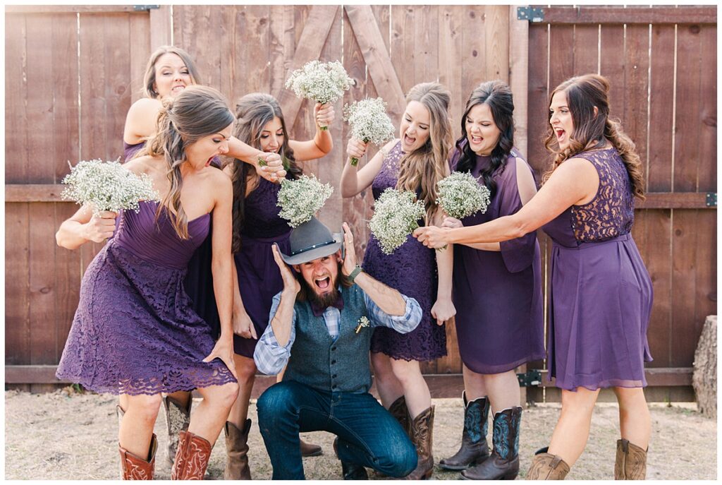 Texas styled bridesmaid in purple dresses and cowboy boots  poses in front of barn door with groom in cowboy hat for bridal party portraits for outdoor Texas styled wedding at Fort Worth Country Memorial Wedding Venue photographed by Dallas wedding photographer Jenny Bui of Picture Bouquet Studio. 
