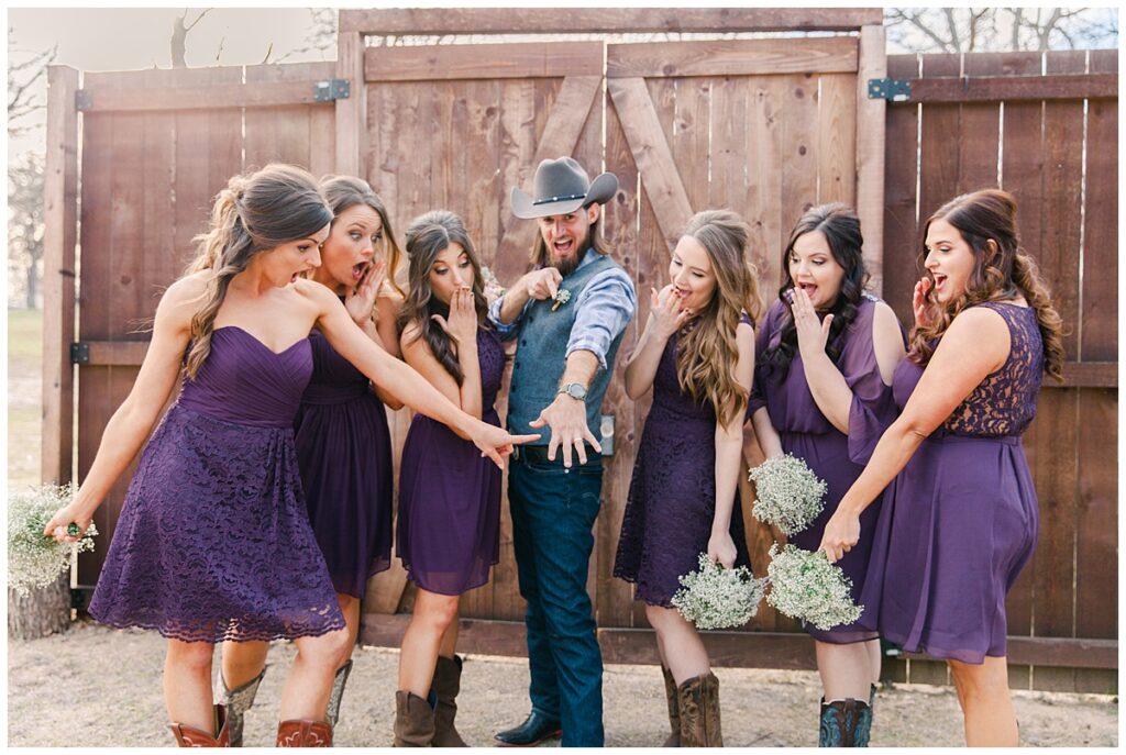 Texas styled bridesmaid in purple dresses and cowboy boots  poses in front of barn door with groom in cowboy hat for bridal party portraits for outdoor Texas styled wedding at Fort Worth Country Memorial Wedding Venue photographed by Dallas wedding photographer Jenny Bui of Picture Bouquet Studio. 