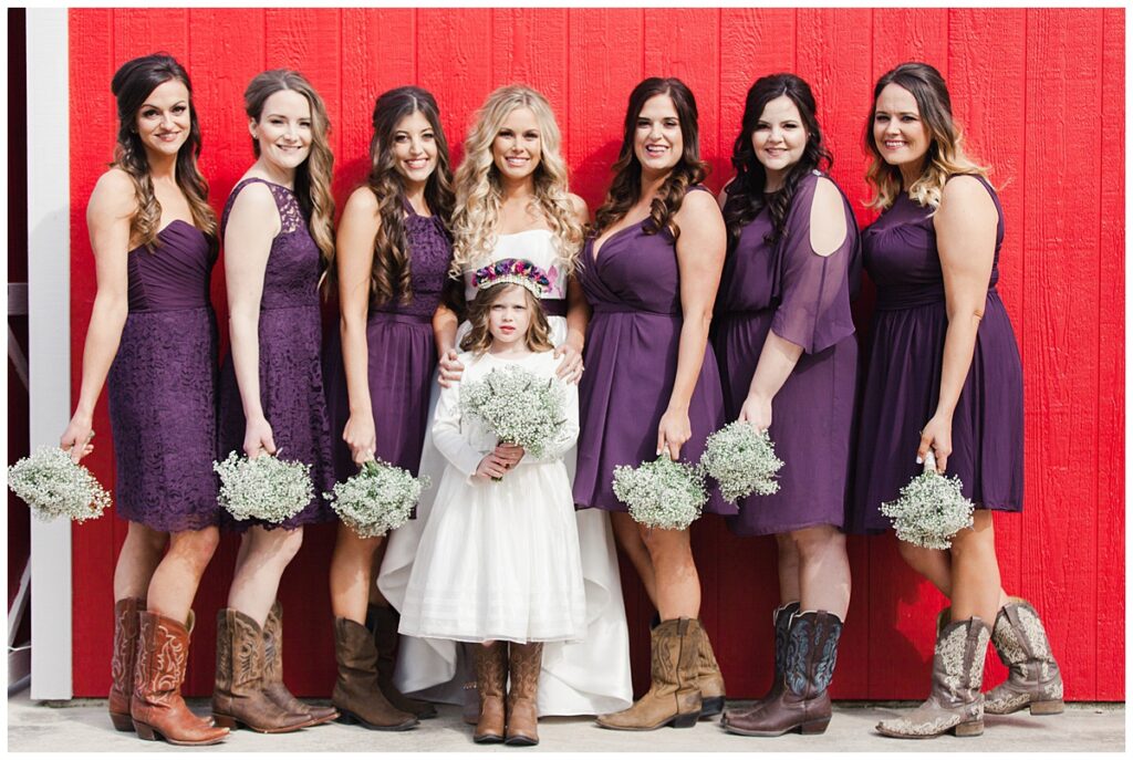 Texas styled bridesmaid poses with bride in purple bridesmaids dresses and baby's breaths bouquet in front of red barn for outdoor Texas styled wedding at Fort Worth Country Memorial Wedding Venue photographed by Dallas wedding photographer Jenny Bui of Picture Bouquet Studio. 