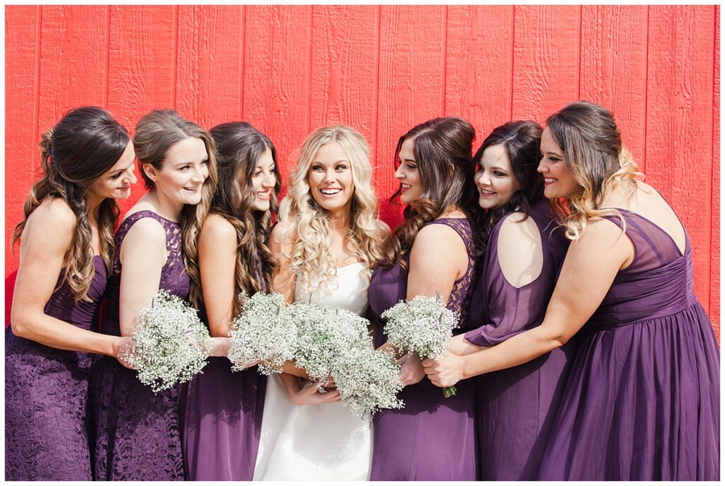 Texas styled bridesmaid poses with bride in purple bridesmaids dresses and baby's breaths bouquet in front of red barn for outdoor Texas styled wedding at Fort Worth Country Memorial Wedding Venue photographed by Dallas wedding photographer Jenny Bui of Picture Bouquet Studio. 