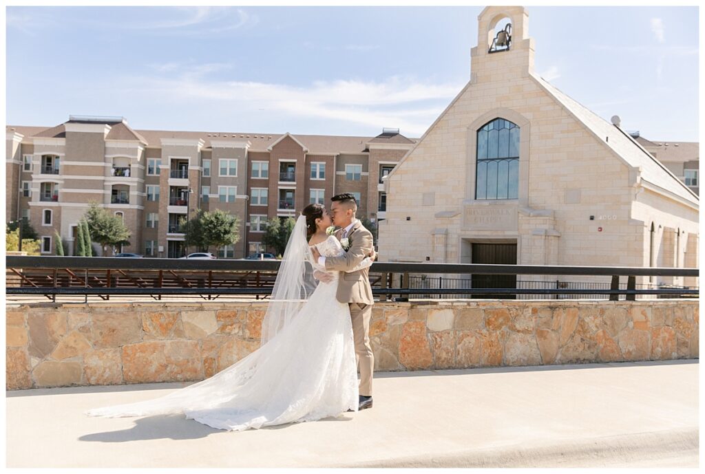 Groom in tan suit and bride in long sleeve lace wedding dress kisses in front of The Riverwalk Chapel during first look at Flower Mound River Walk photographed by Dallas wedding photographer, Jenny Bui of Picture Bouquet Studio.  