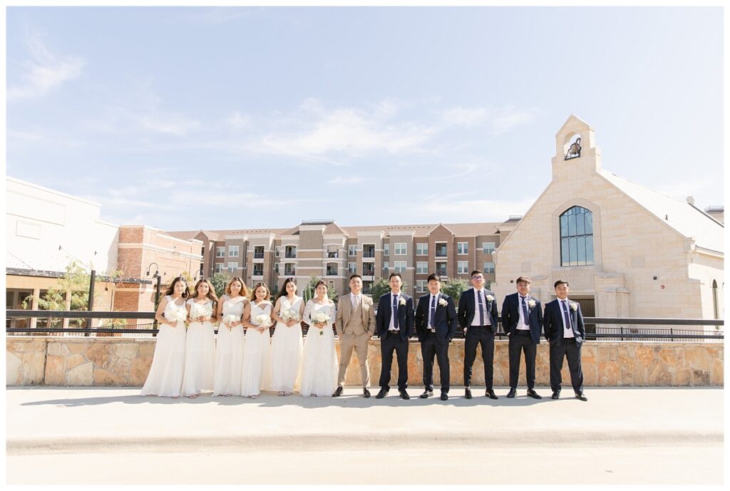 Bride in off shoulder long sleeve white lace wedding dress and groom in tan suit poses with bridesmaid in long white gowns in front of The Riverwalk Chapel during bridal party portraits at Flower Mound River Walk photographed by Dallas wedding photographer, Jenny Bui of Picture Bouquet Studio.   