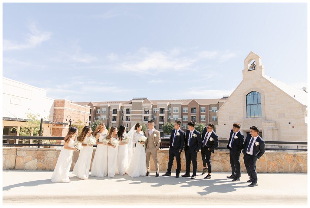 Bridal party looks at bride in off shoulder long sleeve white lace wedding dress and groom in tan suit in front of The Riverwalk Chapel during bridal party portraits at Flower Mound River Walk photographed by Dallas wedding photographer, Jenny Bui of Picture Bouquet Studio.   