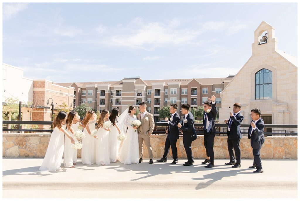 Bridal party looks and cheers at bride in off shoulder long sleeve white lace wedding dress and groom in tan suit in front of The Riverwalk Chapel during bridal party portraits at Flower Mound River Walk photographed by Dallas wedding photographer, Jenny Bui of Picture Bouquet Studio.   