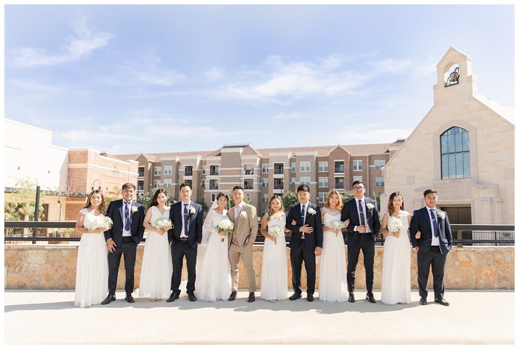 Bridal party poses for camera with bride in off shoulder long sleeve white lace wedding dress and groom in tan suit in front of The Riverwalk Chapel during bridal party portraits at Flower Mound River Walk photographed by Dallas wedding photographer, Jenny Bui of Picture Bouquet Studio.   