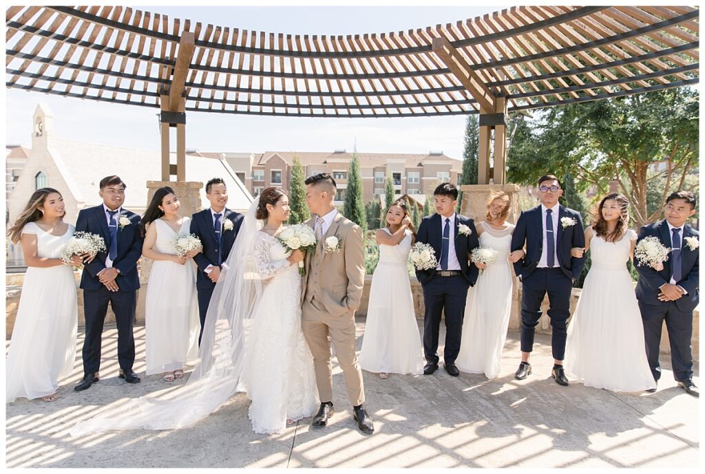 Bridal party looks at bride in off shoulder long sleeve white lace wedding dress and groom in tan suit under awning during bridal party portraits at Flower Mound River Walk photographed by Dallas wedding photographer, Jenny Bui of Picture Bouquet Studio.   