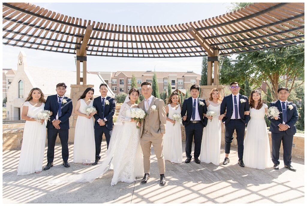 Bridal party poses with bride in off shoulder long sleeve white lace wedding dress and groom in tan suit under wood awning during bridal party portraits at Flower Mound River Walk photographed by Dallas wedding photographer, Jenny Bui of Picture Bouquet Studio.   