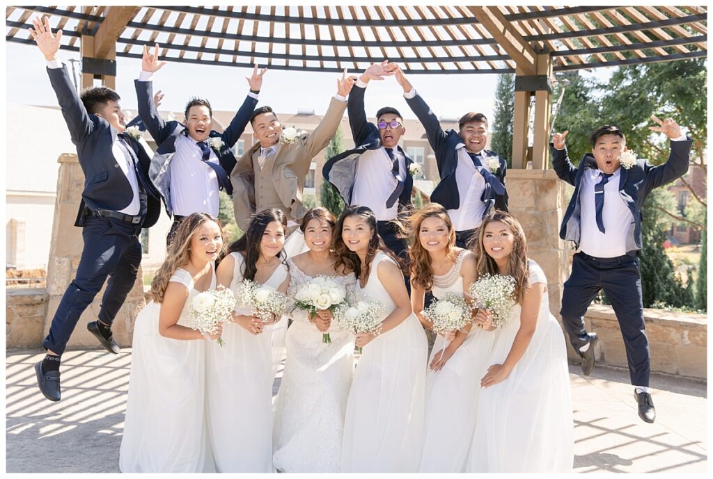 Groomsmen in navy suits and groom in tan suit jumps behind bridesmaids  and bride in white gown under wood awning during bridal party portraits at Flower Mound River Walk photographed by Dallas wedding photographer, Jenny Bui of Picture Bouquet Studio.   