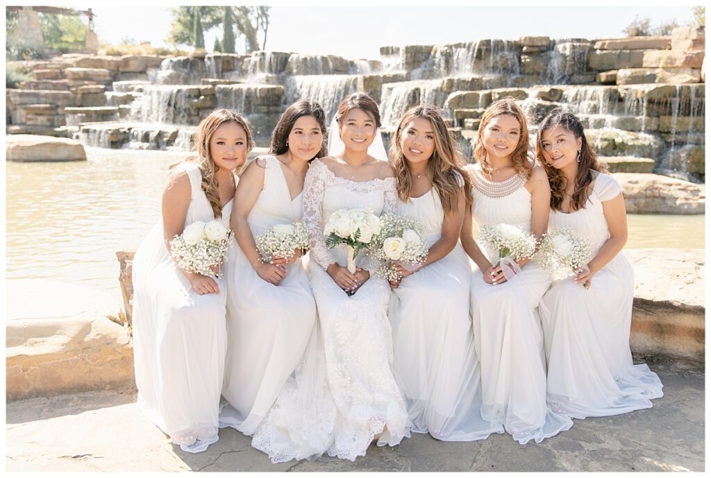 Bride in off shoulder long sleeve white lace wedding gown poses with bridesmaids in long white gowns in front of rock waterfall during bridal party portraits at Flower Mound River Walk photographed by Dallas wedding photographer, Jenny Bui of Picture Bouquet Studio. 