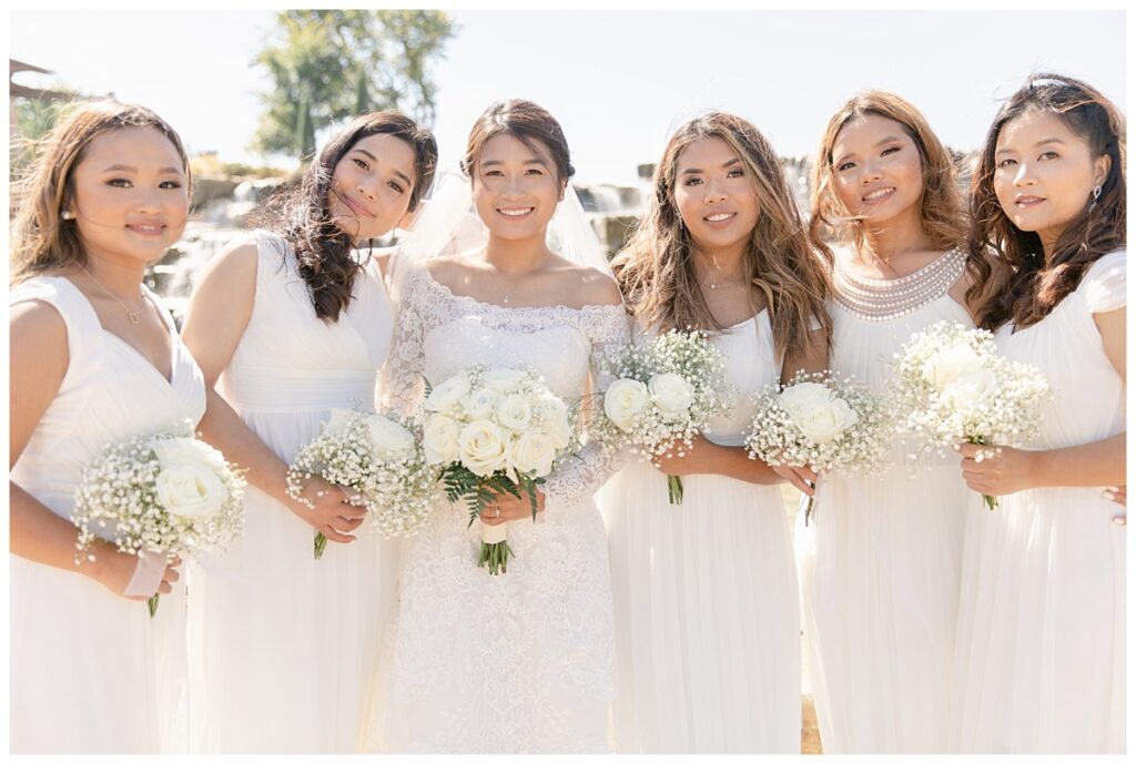 Bridesmaids in long white silk gowns smiles at camera with bride in off shoulder long sleeve white lace wedding gown in front of rock waterfall during bridal party portraits at Flower Mound River Walk photographed by Dallas wedding photographer, Jenny Bui of Picture Bouquet Studio. 
