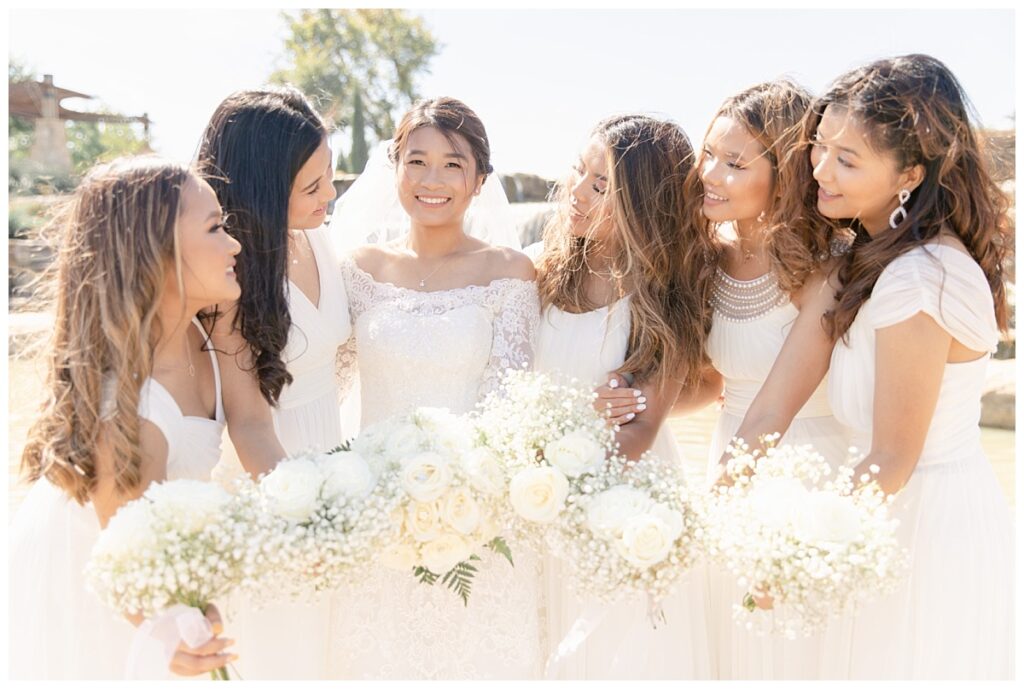 Bridesmaids in long white silk gowns smiles at bride in off shoulder long sleeve white lace wedding gown in front of rock waterfall during bridal party portraits at Flower Mound River Walk photographed by Dallas wedding photographer, Jenny Bui of Picture Bouquet Studio. 