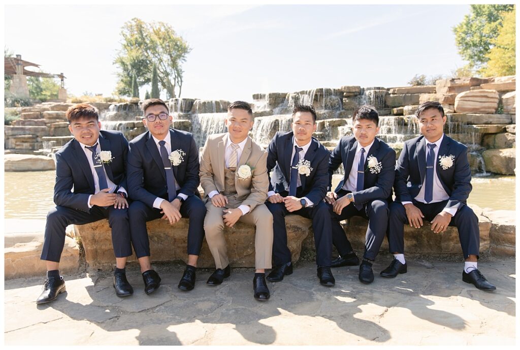 Groomsmen in navy suits poses with groom in tan suit sitting in front of rock waterfall during bridal party portraits at Flower Mound River Walk photographed by Dallas wedding photographer, Jenny Bui of Picture Bouquet Studio. 