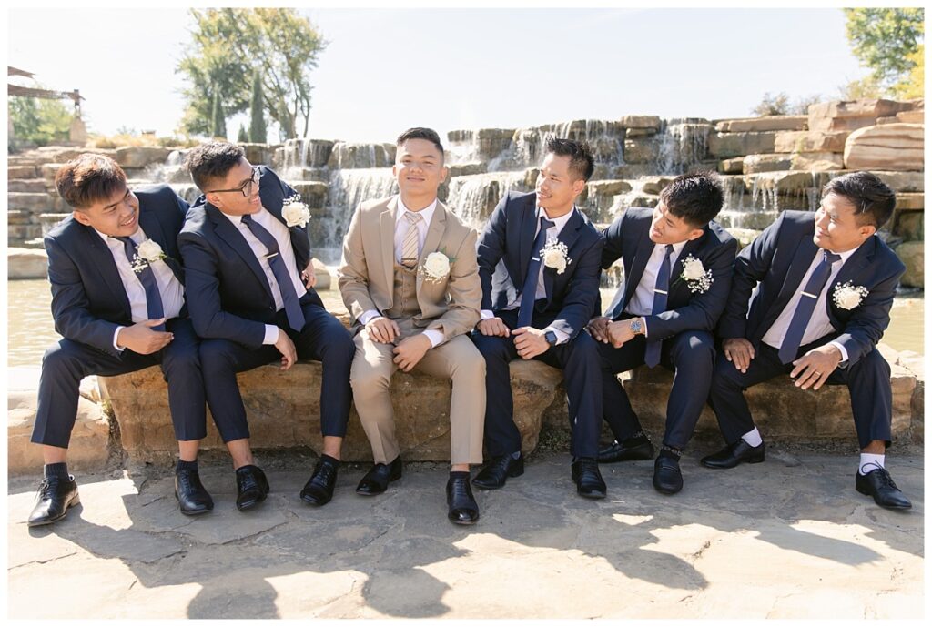 Groomsmen in navy suits smiles at  groom in tan suit sitting in front of rock waterfall during bridal party portraits at Flower Mound River Walk photographed by Dallas wedding photographer, Jenny Bui of Picture Bouquet Studio. 