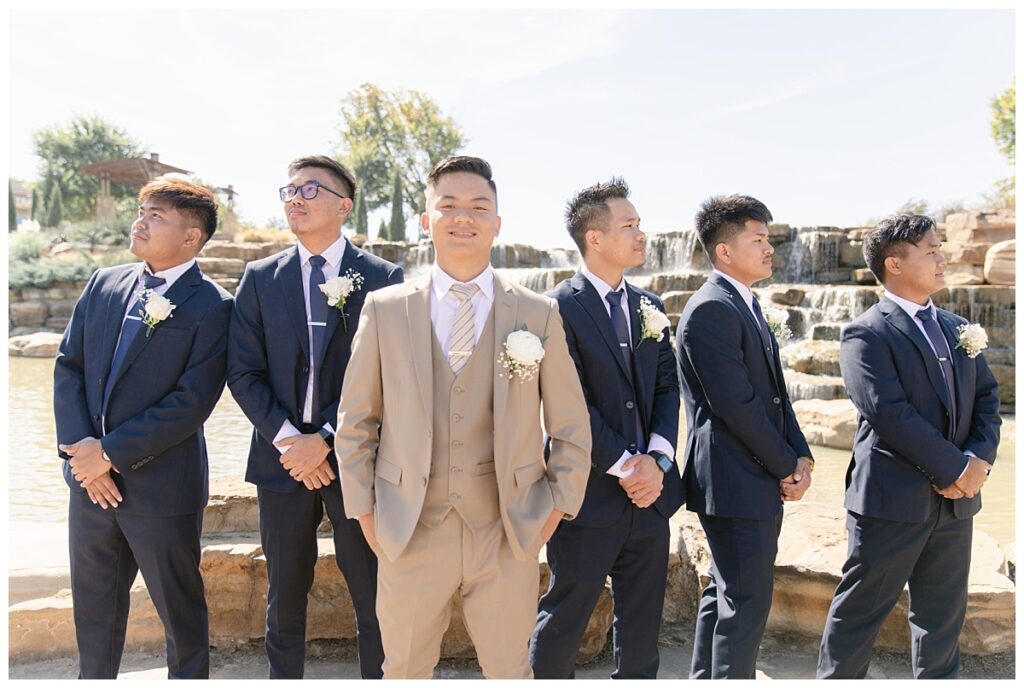 Groomsmen in navy suits poses with groom in tan suit standing in front of rock waterfall during bridal party portraits at Flower Mound River Walk photographed by Dallas wedding photographer, Jenny Bui of Picture Bouquet Studio. 