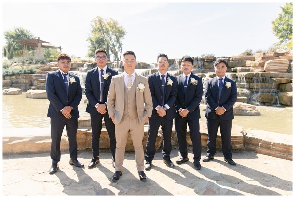 Groomsmen in navy suits poses with groom in tan suit standing in front of rock waterfall during bridal party portraits at Flower Mound River Walk photographed by Dallas wedding photographer, Jenny Bui of Picture Bouquet Studio. 