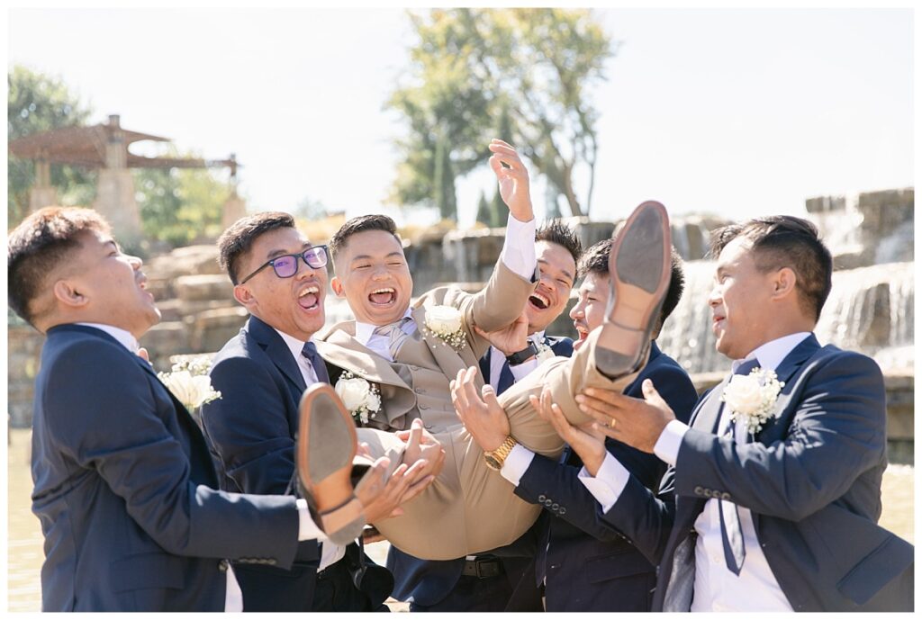 Groomsmen in navy suits throws groom in tan suit in the air in front of rock waterfall during bridal party portraits at Flower Mound River Walk photographed by Dallas wedding photographer, Jenny Bui of Picture Bouquet Studio. 