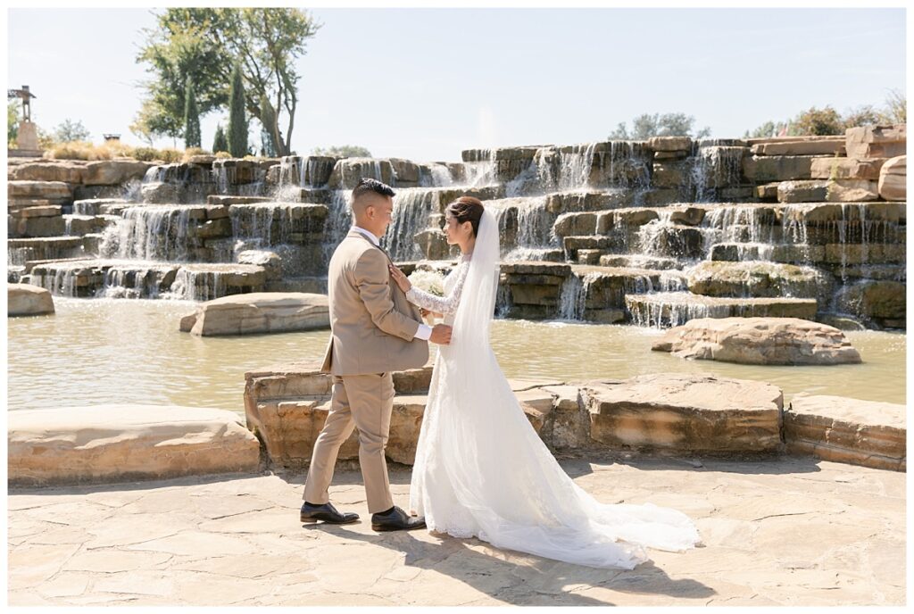 Groom in tan suit walks up to bride in off shoulder long sleeve lace wedding dress and long veil with white bouquet in front of rock waterfall during bridal party portraits at Flower Mound River Walk photographed by Dallas wedding photographer, Jenny Bui of Picture Bouquet Studio. 