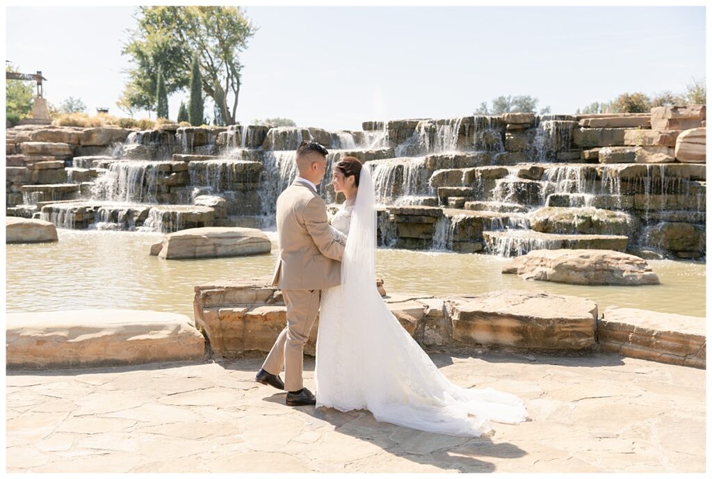 Groom in tan suit hugs bride in off shoulder long sleeve lace wedding dress and long veil with white bouquet in front of rock waterfall during bridal party portraits at Flower Mound River Walk photographed by Dallas wedding photographer, Jenny Bui of Picture Bouquet Studio. 