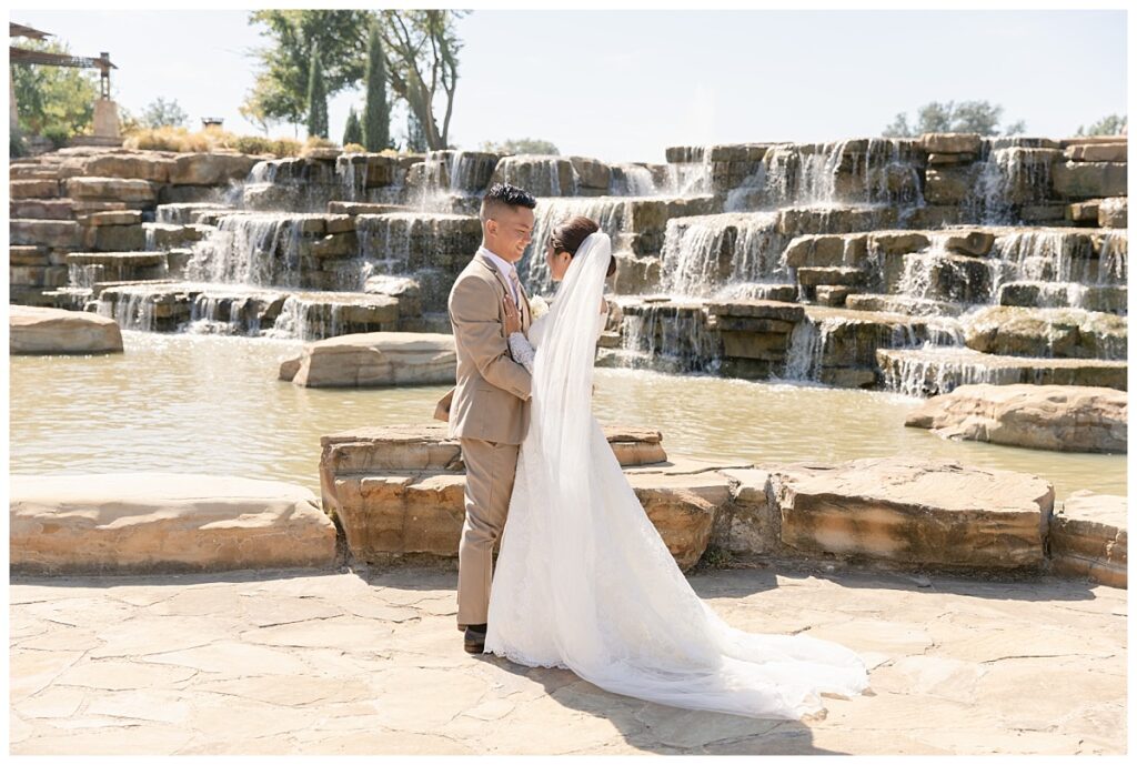 Groom in tan suit hugs and looks at bride in off shoulder long sleeve lace wedding dress and long veil with white bouquet in front of rock waterfall during bridal party portraits at Flower Mound River Walk photographed by Dallas wedding photographer, Jenny Bui of Picture Bouquet Studio. 