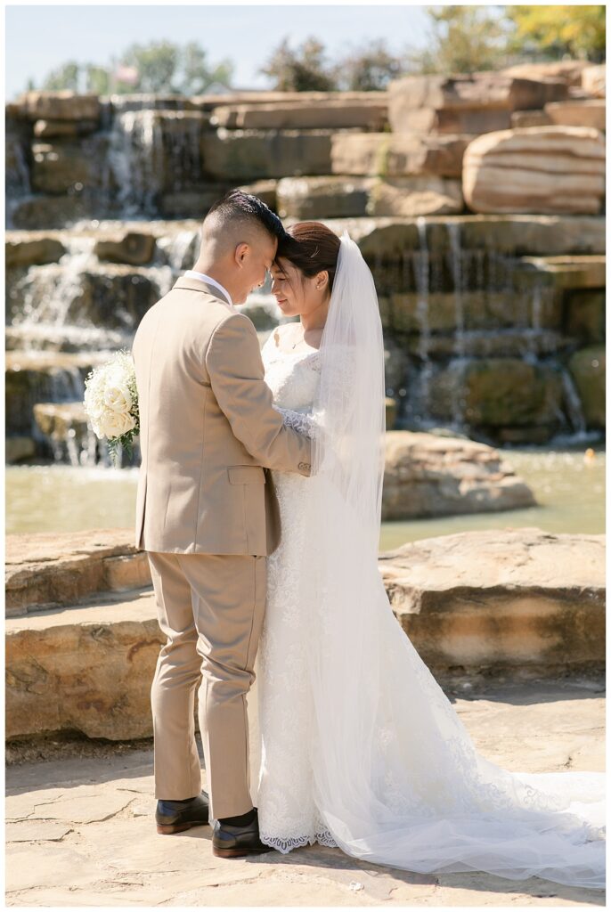 Groom in tan suit presses forehead together with bride in off shoulder long sleeve lace wedding dress and long veil with white bouquet in front of rock waterfall during bridal party portraits at Flower Mound River Walk photographed by Dallas wedding photographer, Jenny Bui of Picture Bouquet Studio. 