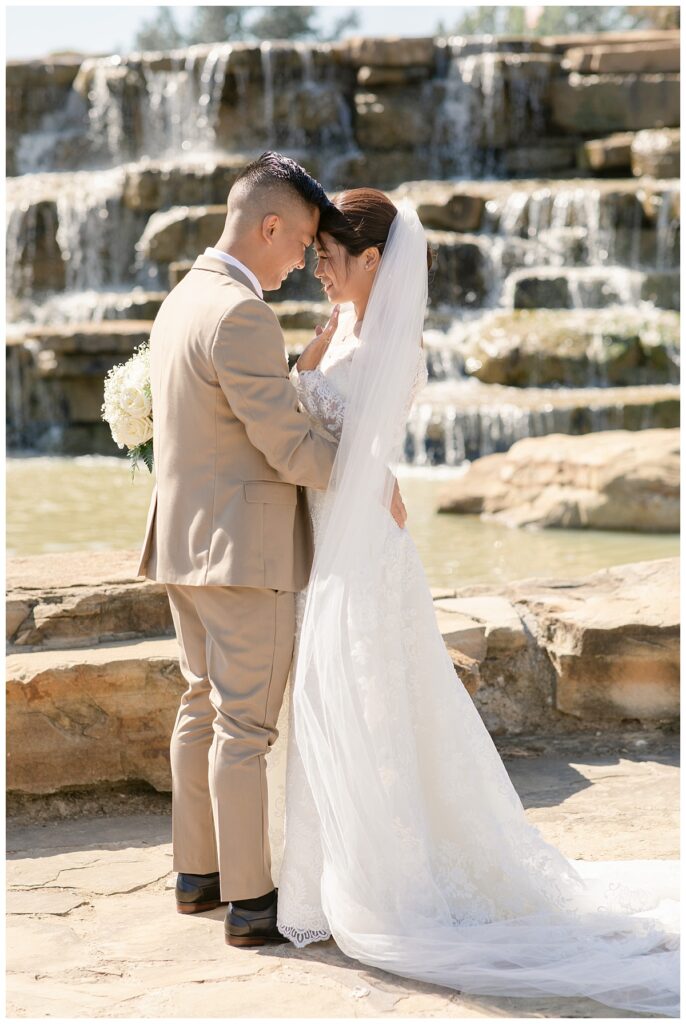 Groom in tan suit presses forehead together with bride and smiles in off shoulder long sleeve lace wedding dress and long veil with white bouquet in front of rock waterfall during bridal party portraits at Flower Mound River Walk photographed by Dallas wedding photographer, Jenny Bui of Picture Bouquet Studio. 