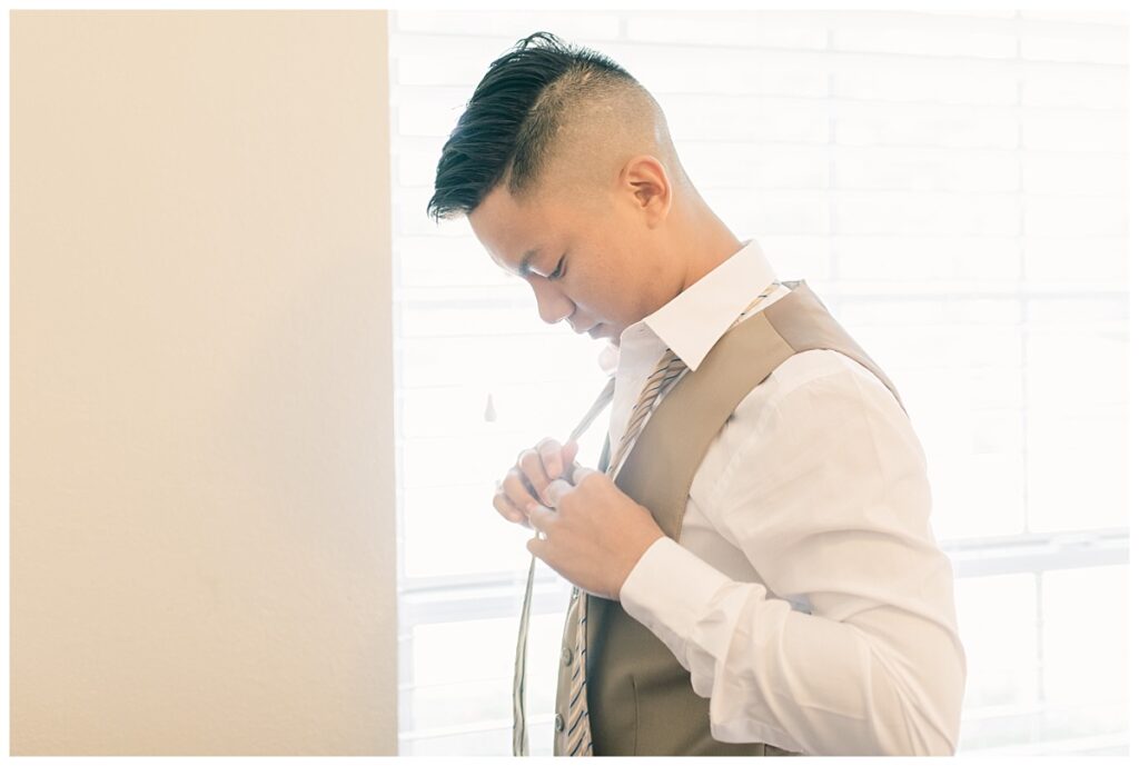 Groom in tan suit putting on tie during bridal prep photographed by Dallas wedding photographer, Jenny Bui of Picture Bouquet Studio. 
