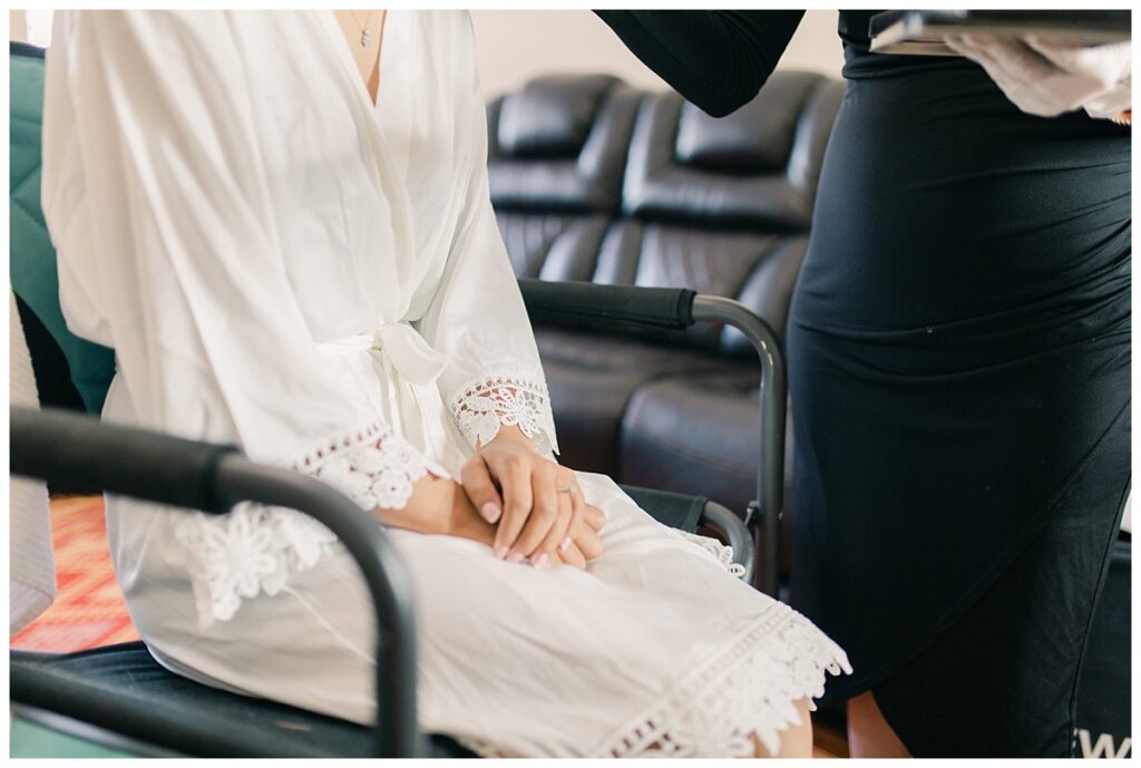 Bride with hand on lap in white silk robe with embroidered appliques sitting in chair getting make up done photographed by Dallas wedding photographer, Jenny Bui of Picture Bouquet Studio. 
