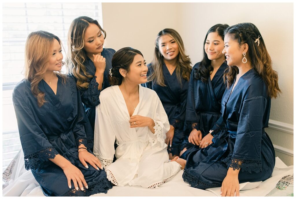 Bride in white silk robe and bridesmaids in navy silk robe smiles and laughs during bridal prep photographed by Dallas wedding photographer, Jenny Bui of Picture Bouquet Studio. 