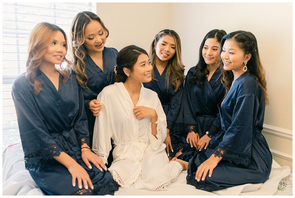 Bride in white silk robe and bridesmaids in navy silk robe smiles at one another during bridal prep photographed by Dallas wedding photographer, Jenny Bui of Picture Bouquet Studio. 