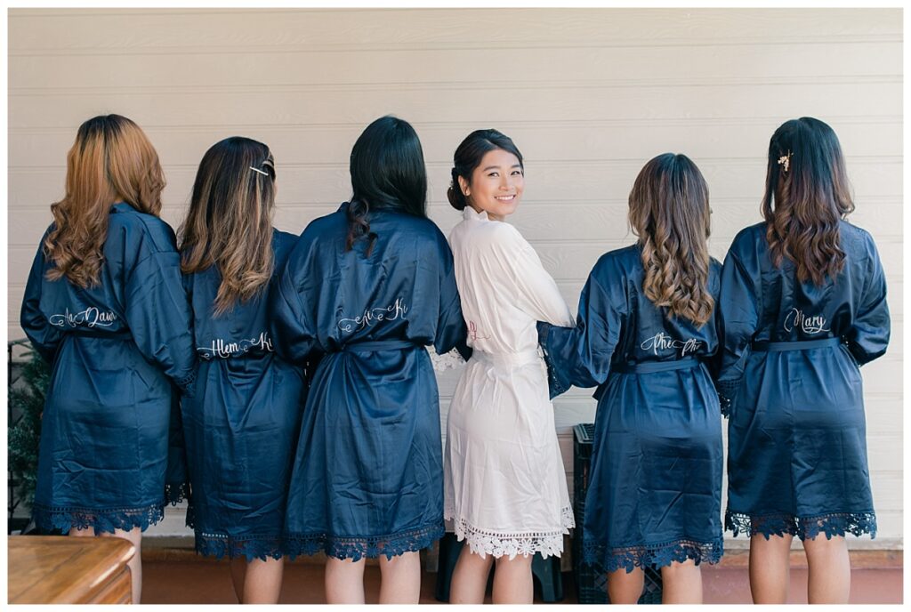 Bride in white silk robe links arm with bridesmaids in navy silk robes smiles and looks back at camera during bridal prep photographed by Dallas wedding photographer, Jenny Bui of Picture Bouquet Studio.  