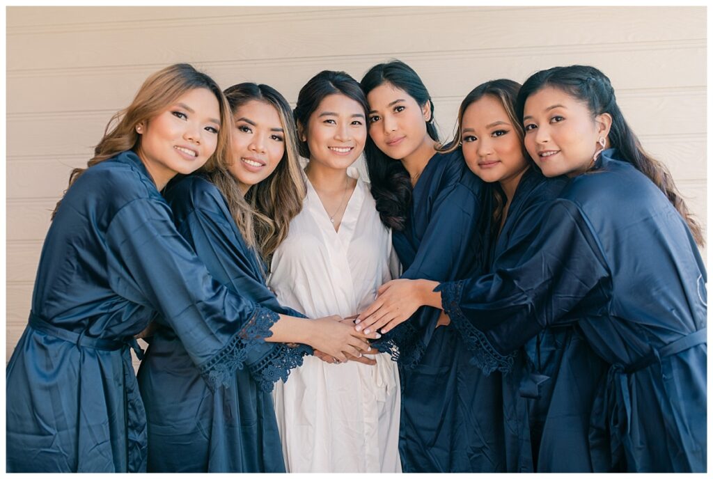 Bride in white silk robe and bridesmaids in navy silk robe hugs each other and smiles at camera during bridal prep photographed by Dallas wedding photographer, Jenny Bui of Picture Bouquet Studio. 