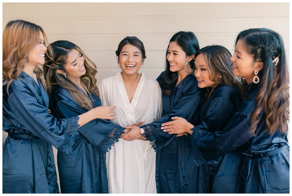 Bride in white silk robe and bridesmaids in navy silk robe hugs and laughs together during bridal prep photographed by Dallas wedding photographer, Jenny Bui of Picture Bouquet Studio. 