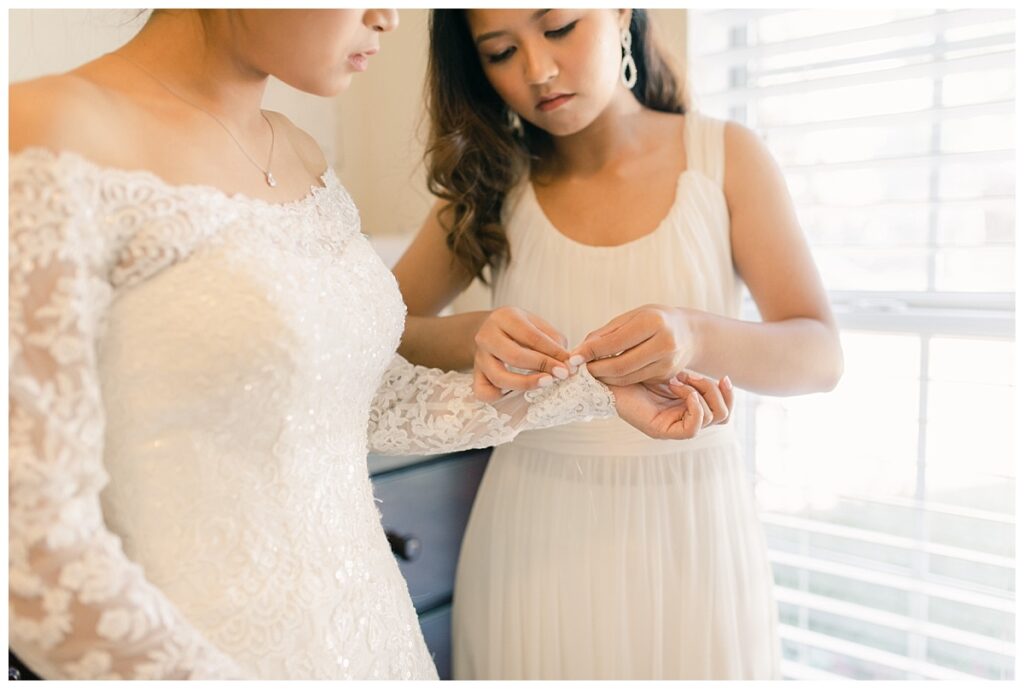 Bridesmaid in white silk dress helps bride in white lace long sleeve wedding dress button up bride's sleeve during bridal prep photographed by Dallas wedding photographer, Jenny Bui of Picture Bouquet Studio. 
