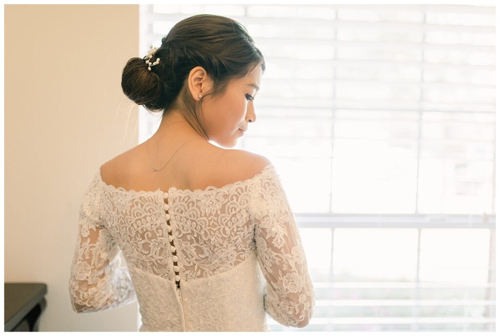 Bride in white off shoulder long sleeve lace wedding dress looks down for detail shot of back during bridal prep photographed by Dallas wedding photographer, Jenny Bui of Picture Bouquet Studio. 