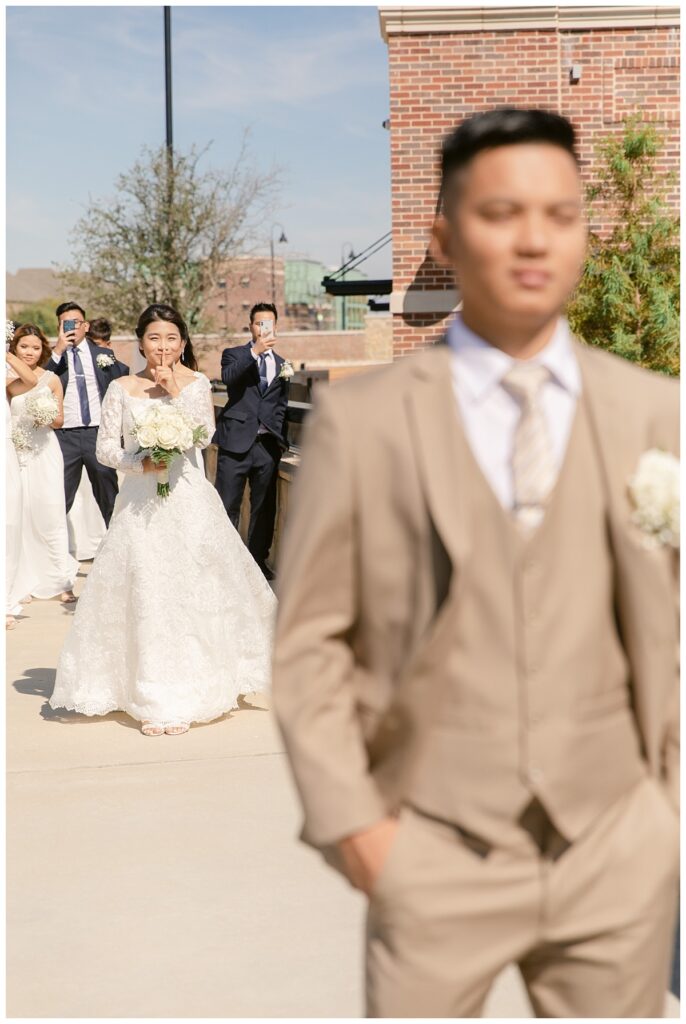 Bride in white off shoulder long sleeve lace wedding dress walks up behind groom in tan suit during first look at Flower Mound River Walk photographed by Dallas wedding photographer, Jenny Bui of Picture Bouquet Studio.  