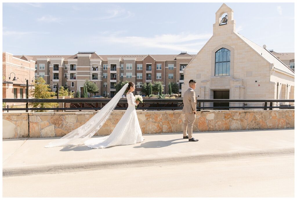 Bride in off shoulder lace long sleeve wedding dress walking up to groom in tan suit in front of The Riverwalk Chapel during first look at Flower Mound River Walk photographed by Dallas wedding photographer, Jenny Bui of Picture Bouquet Studio.  
