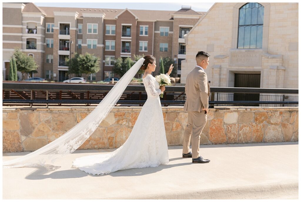 Bride in off shoulder lace long sleeve wedding dress walking up to groom in tan suit in front of The Riverwalk Chapel during first look at Flower Mound River Walk photographed by Dallas wedding photographer, Jenny Bui of Picture Bouquet Studio.  