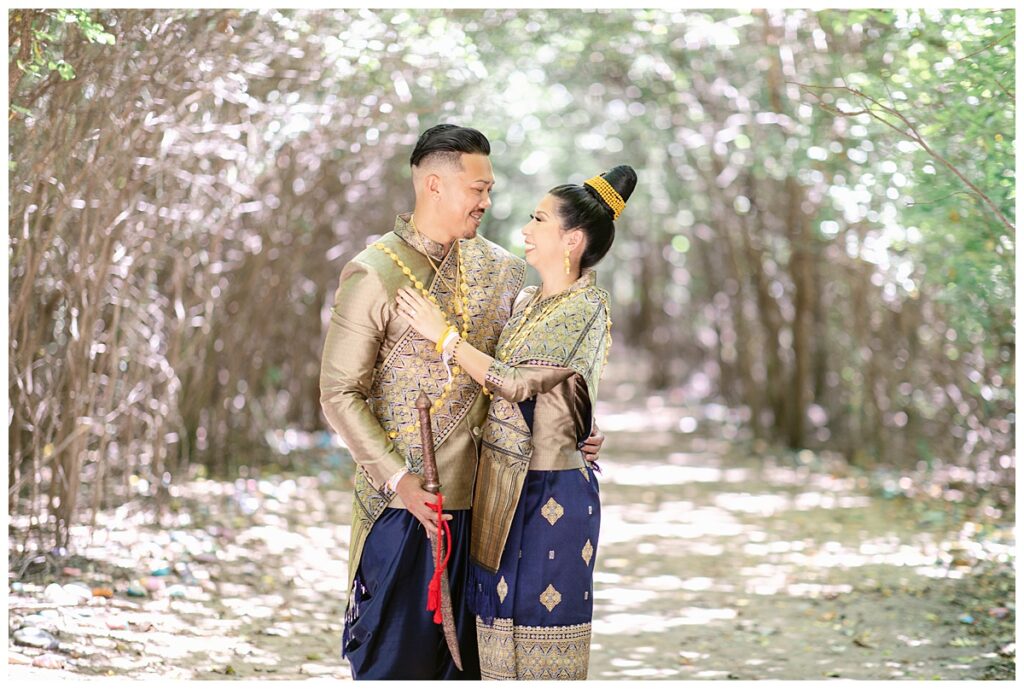 Bride and groom in Laotian navy and gold traditional wedding attire hugs and smiles at one another for Laotian wedding portrait session in tunnel of trees at North Richland HIll's park for Jenny Bui of Picture Bouquet Studio, a wedding photography studio serving the Dallas and surrounding areas. 