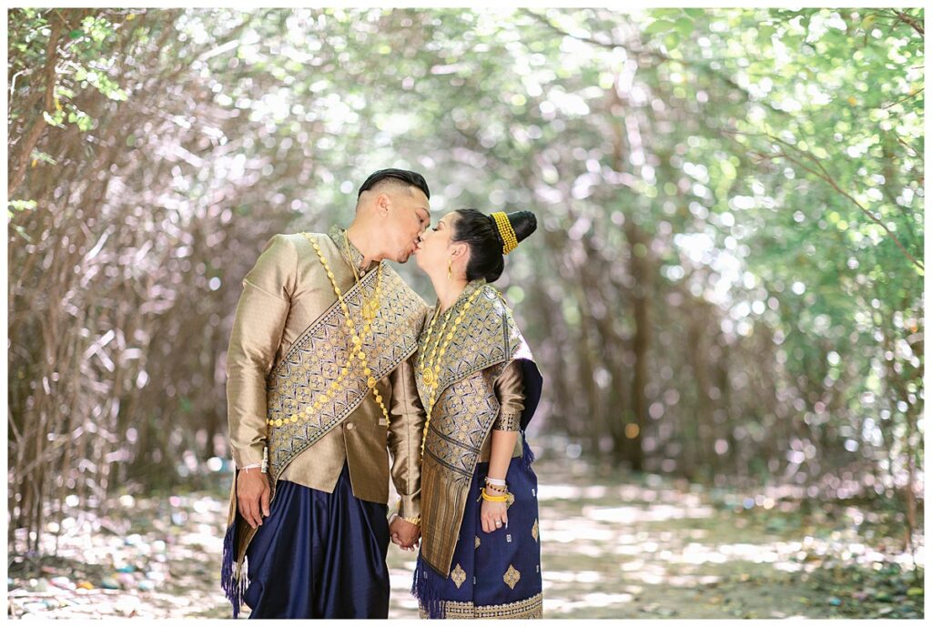 Bride and groom in Laotian navy and gold traditional wedding attire leans in for kiss for Laotian wedding portrait session in tunnel of trees at North Richland HIll's park for Jenny Bui of Picture Bouquet Studio, a wedding photography studio serving the Dallas and surrounding areas. 
