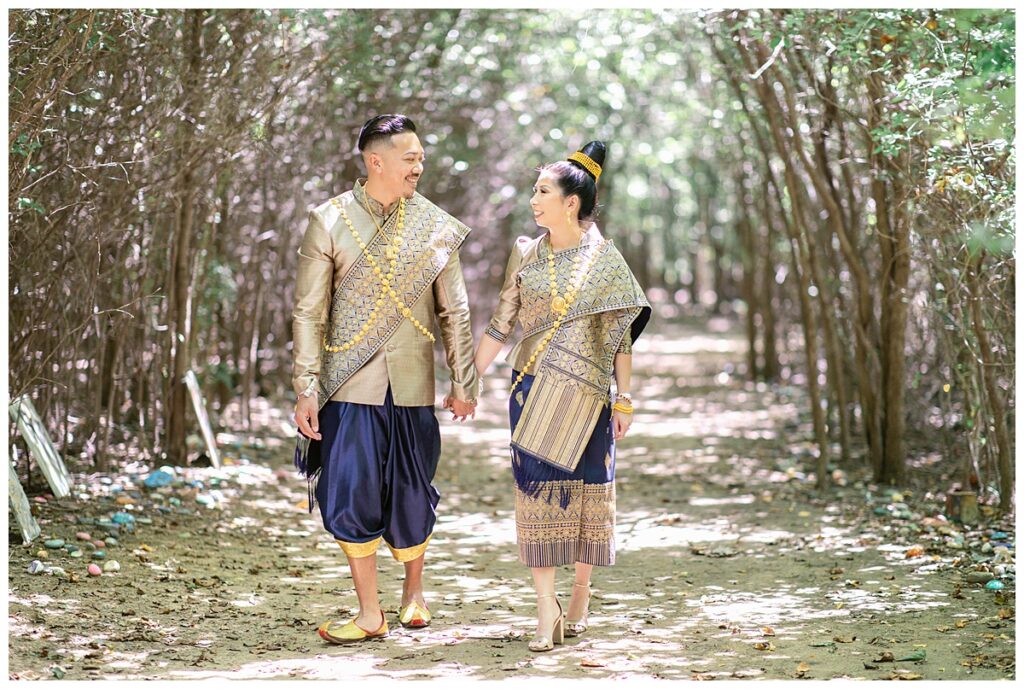Bride and groom in Laotian navy and gold traditional wedding attire holds hands and walks towards camera for Laotian wedding portrait session in tunnel of trees at North Richland HIll's park for Jenny Bui of Picture Bouquet Studio, a wedding photography studio serving the Dallas and surrounding areas. 