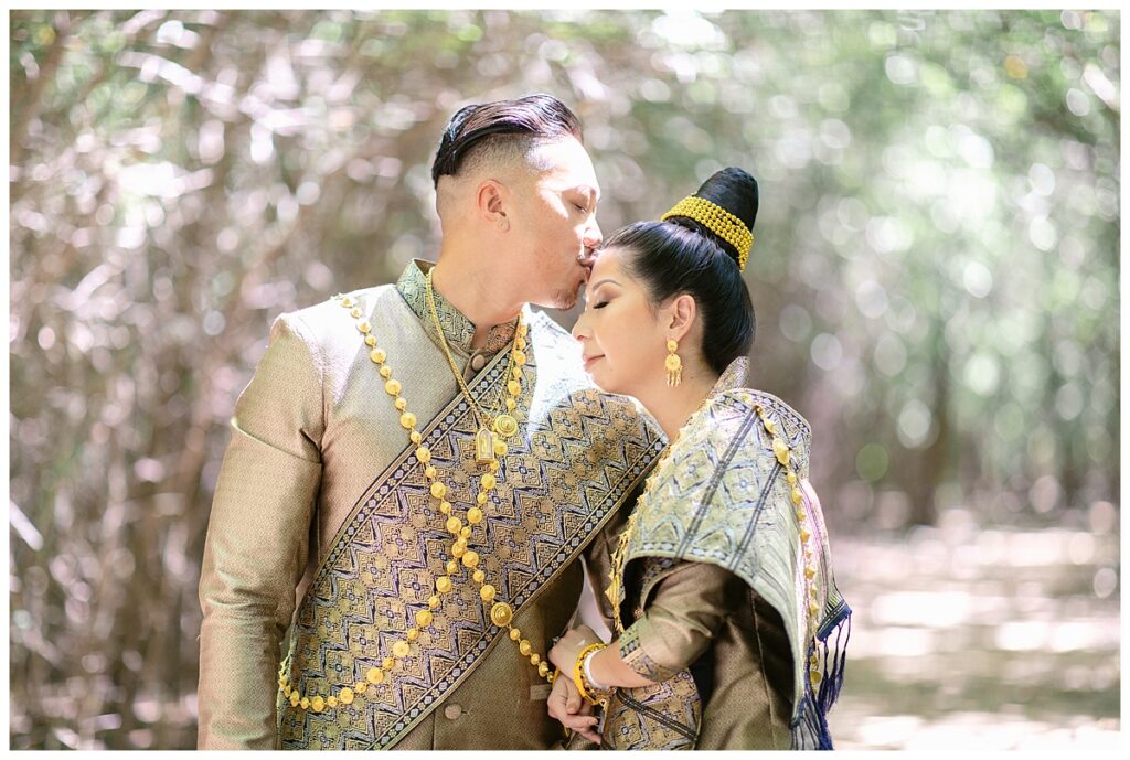 Groom in Laotian navy and gold traditional wedding attire kisses bride on forehead for Laotian wedding portrait session in tunnel of trees at North Richland HIll's park for Jenny Bui of Picture Bouquet Studio, a wedding photography studio serving the Dallas and surrounding areas. 