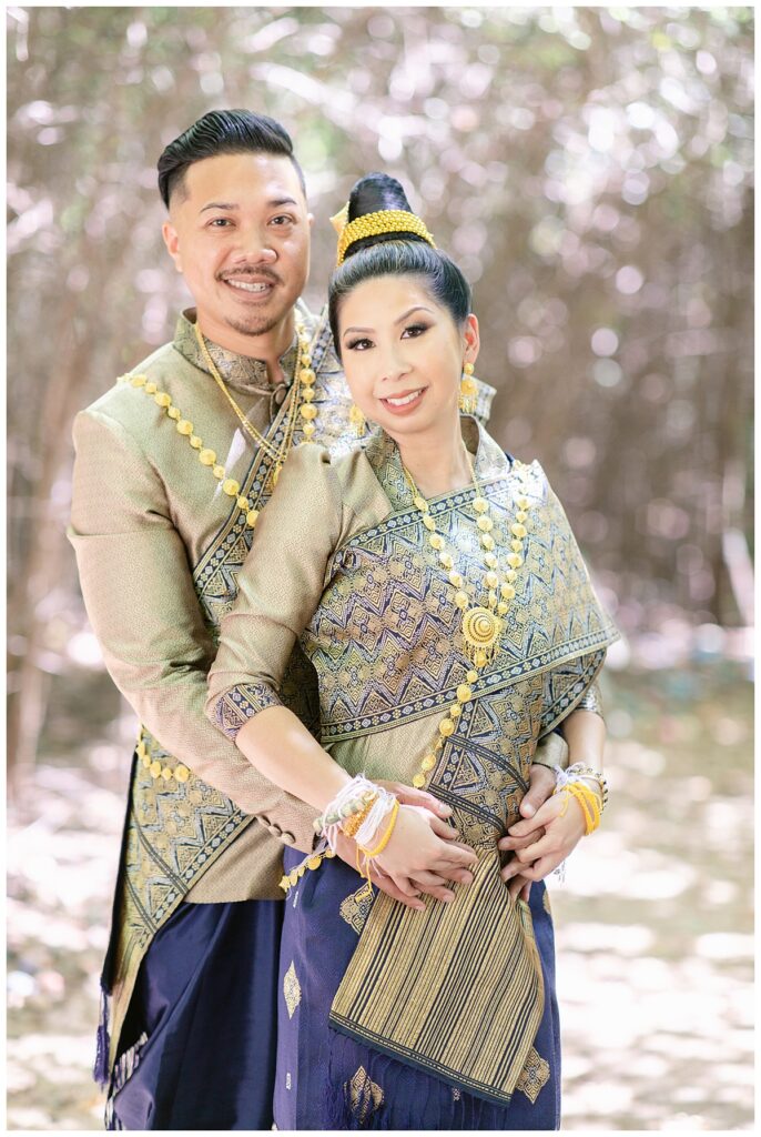 Groom in Laotian navy and gold traditional wedding attire wraps hands around bride's waist for Laotian wedding portrait session in tunnel of trees at North Richland HIll's park for Jenny Bui of Picture Bouquet Studio, a wedding photography studio serving the Dallas and surrounding areas. 